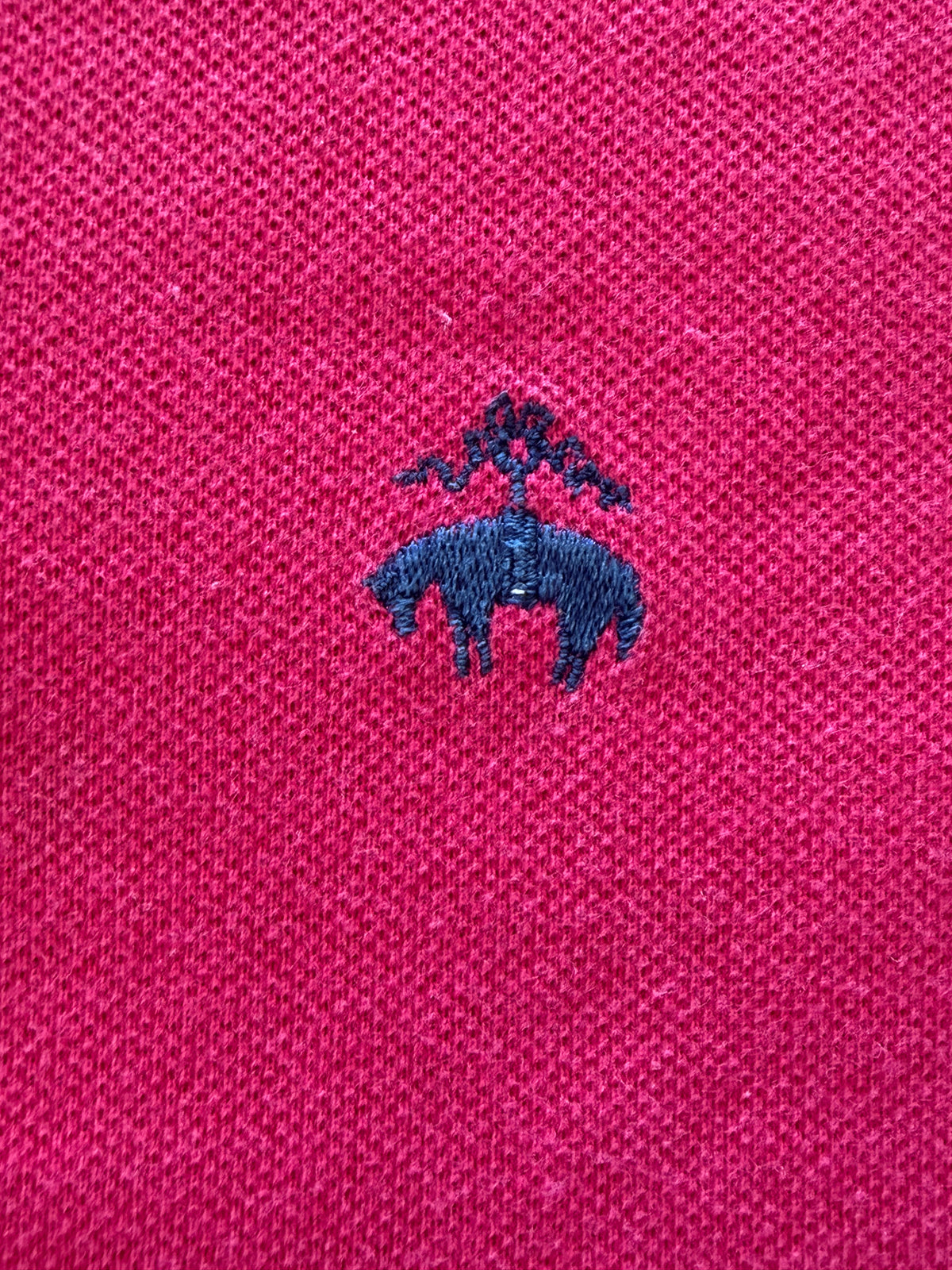 Close-up detail of the navy blue embroidered Brooks Brothers Golden Fleece sheep logo on magenta cotton pique fabric