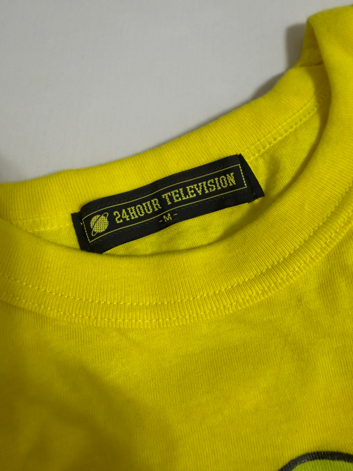 Close-up detail of the black neck label on the yellow Yoshitaka Amano x 24 Hour Television 2014 charity t-shirt, showing the white "24HOUR TELEVISION" text, globe logo, and size "-M-".