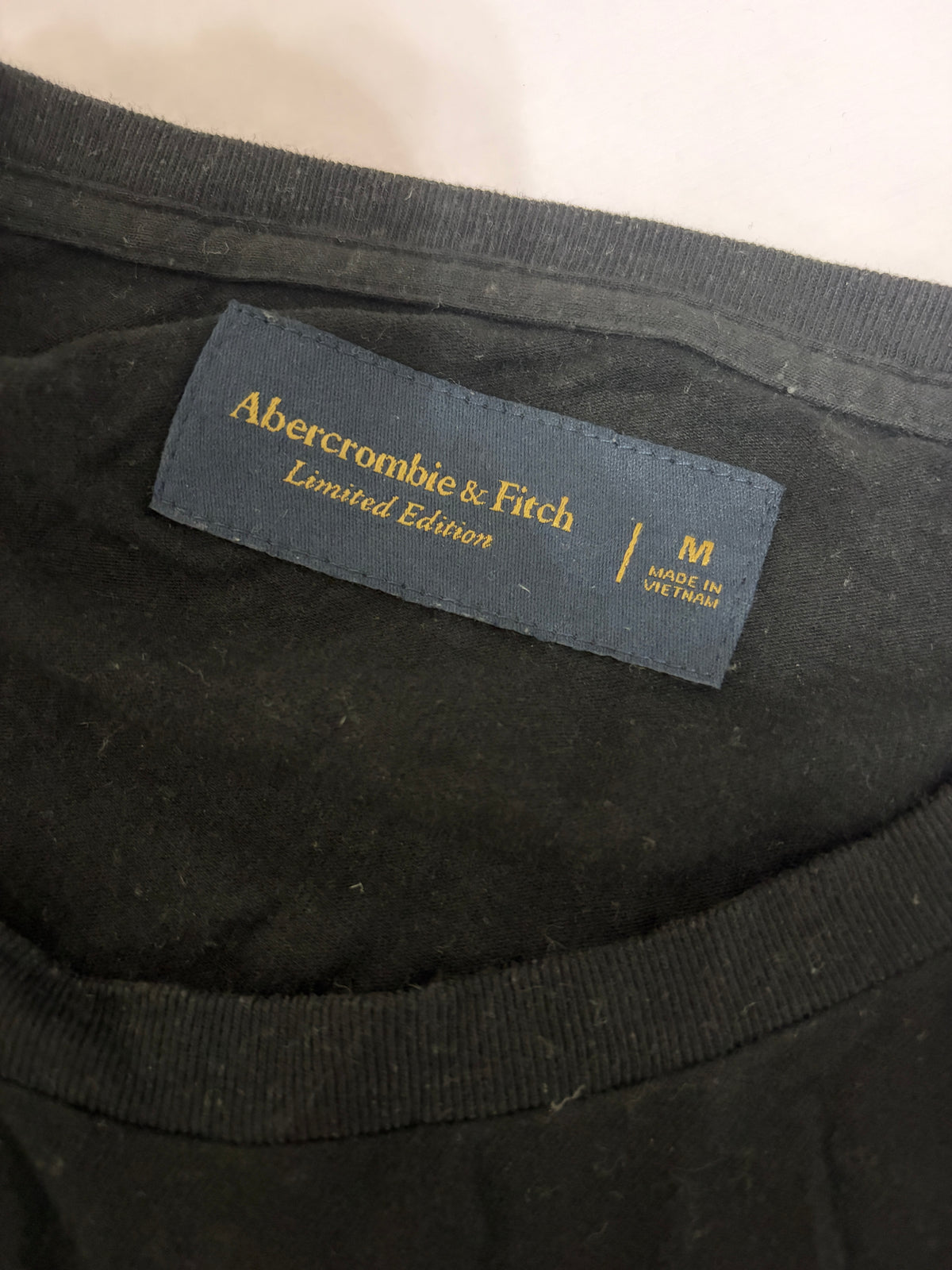 Close-up detail of the navy blue Abercrombie & Fitch Limited Edition woven neck tag and size M label on a black t-shirt