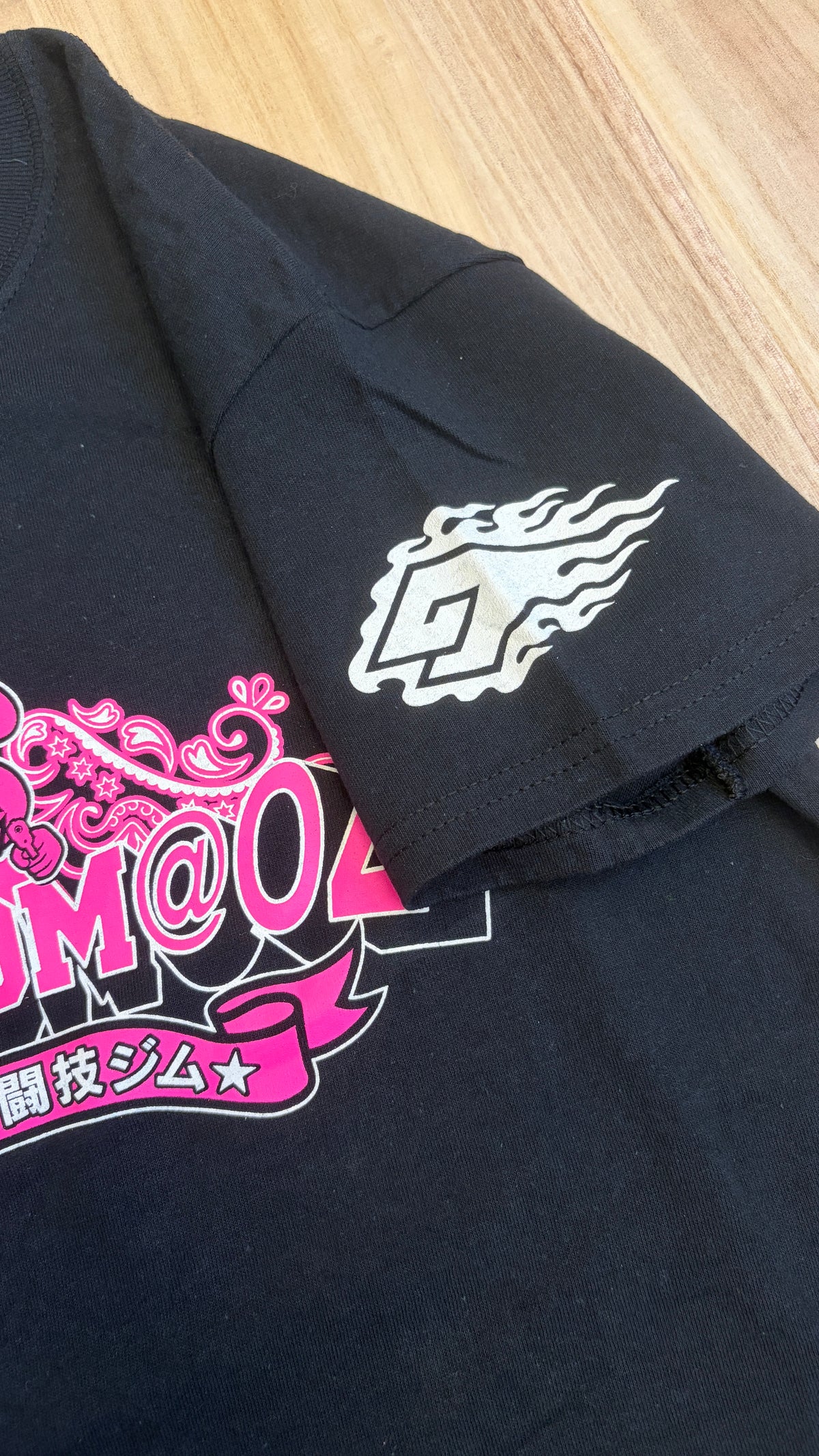 Close-up view of the left sleeve of a black graphic t-shirt featuring a white stylized flaming G logo print alongside pink Japanese text on the chest