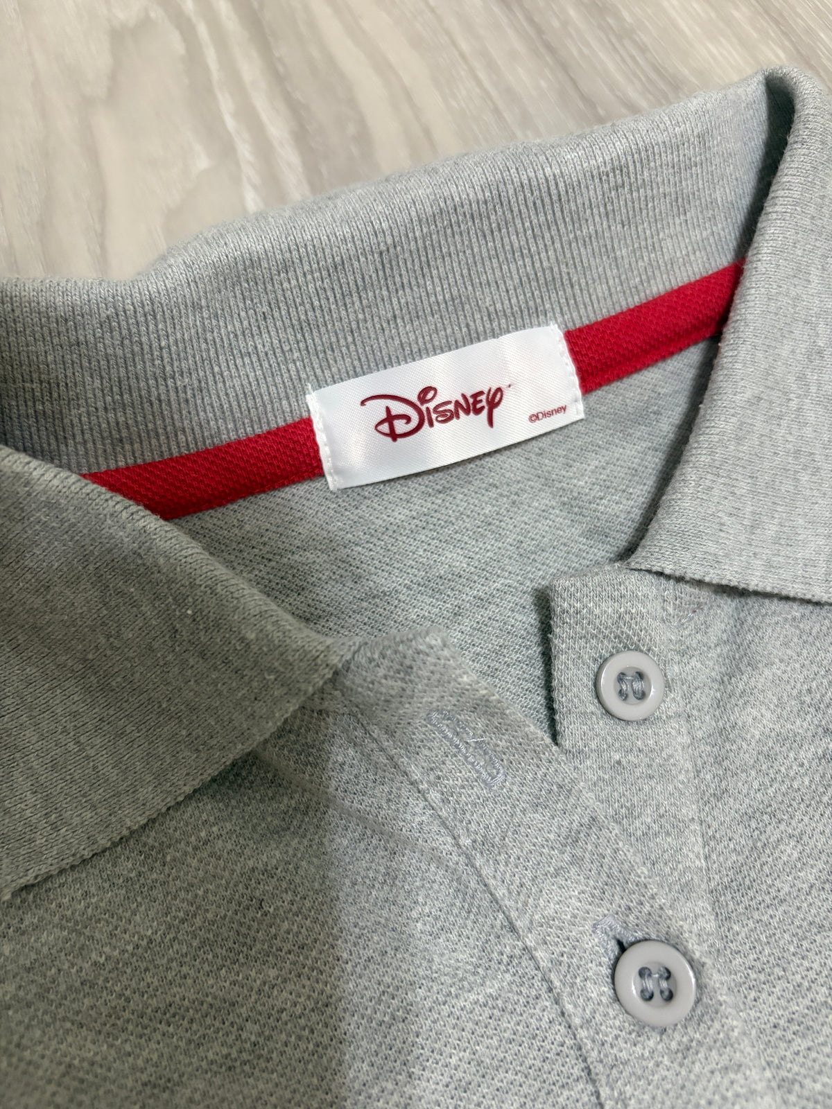 Close-up detail of the white woven Disney brand label stitched on red neck tape inside the collar of a heather grey polo shirt