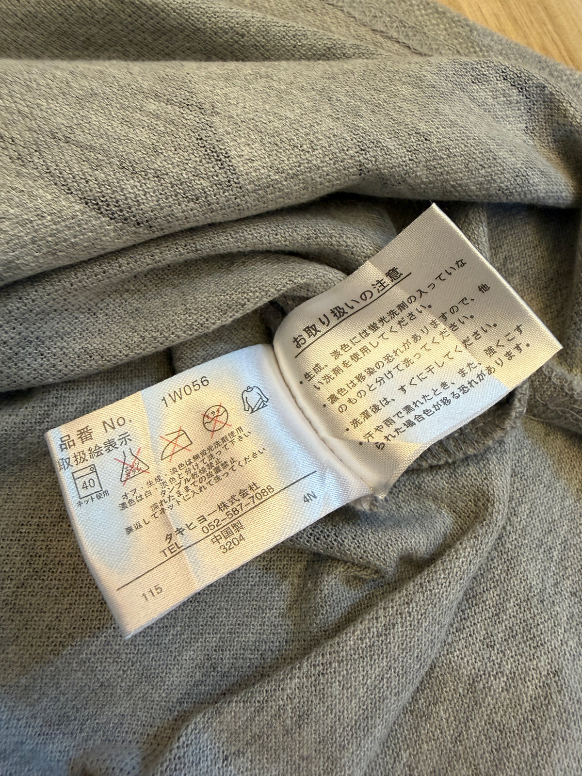 Close-up view of the inner white Japanese care tag on a grey polo shirt displaying item number 1W056, laundry symbols, and manufacturer Takihyo Co., Ltd. details