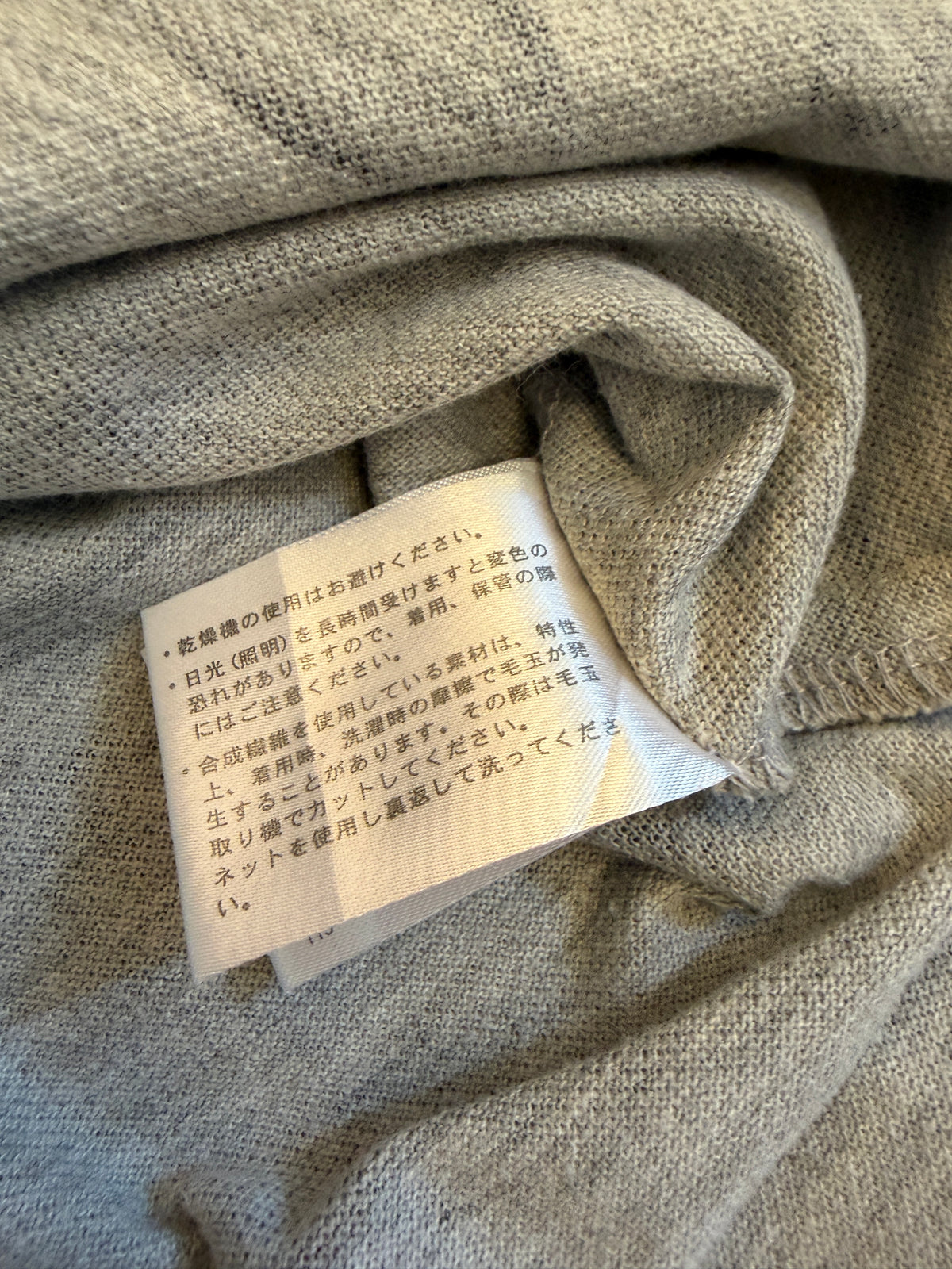 Close-up view of the back side of an inner care tag on a heather grey knit garment featuring Japanese text warnings about pilling, tumble drying, and light exposure