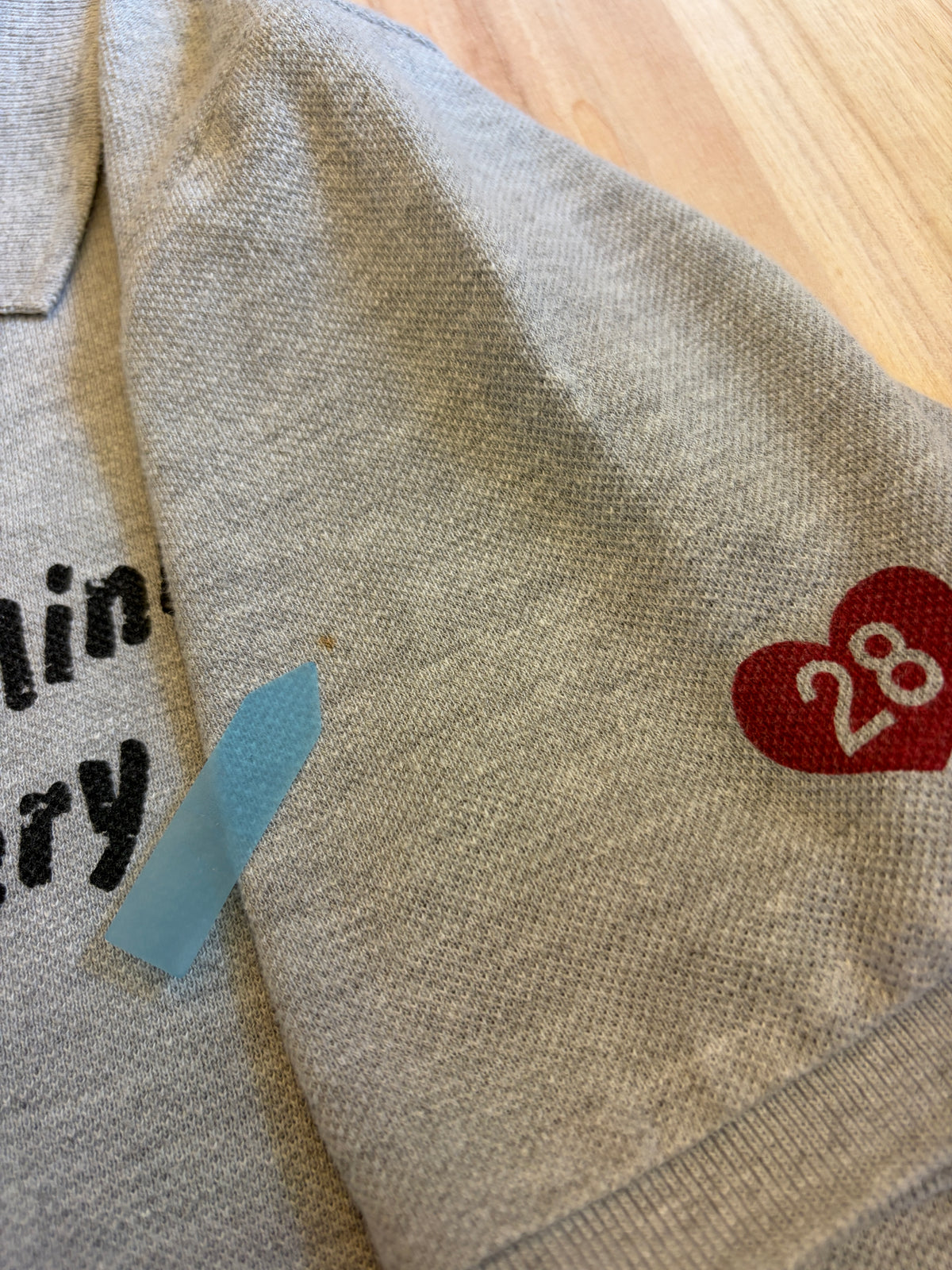 Close-up view of a grey Disney Minnie Mouse polo shirt sleeve showing a small brown stain defect pointed out by a blue sticker.