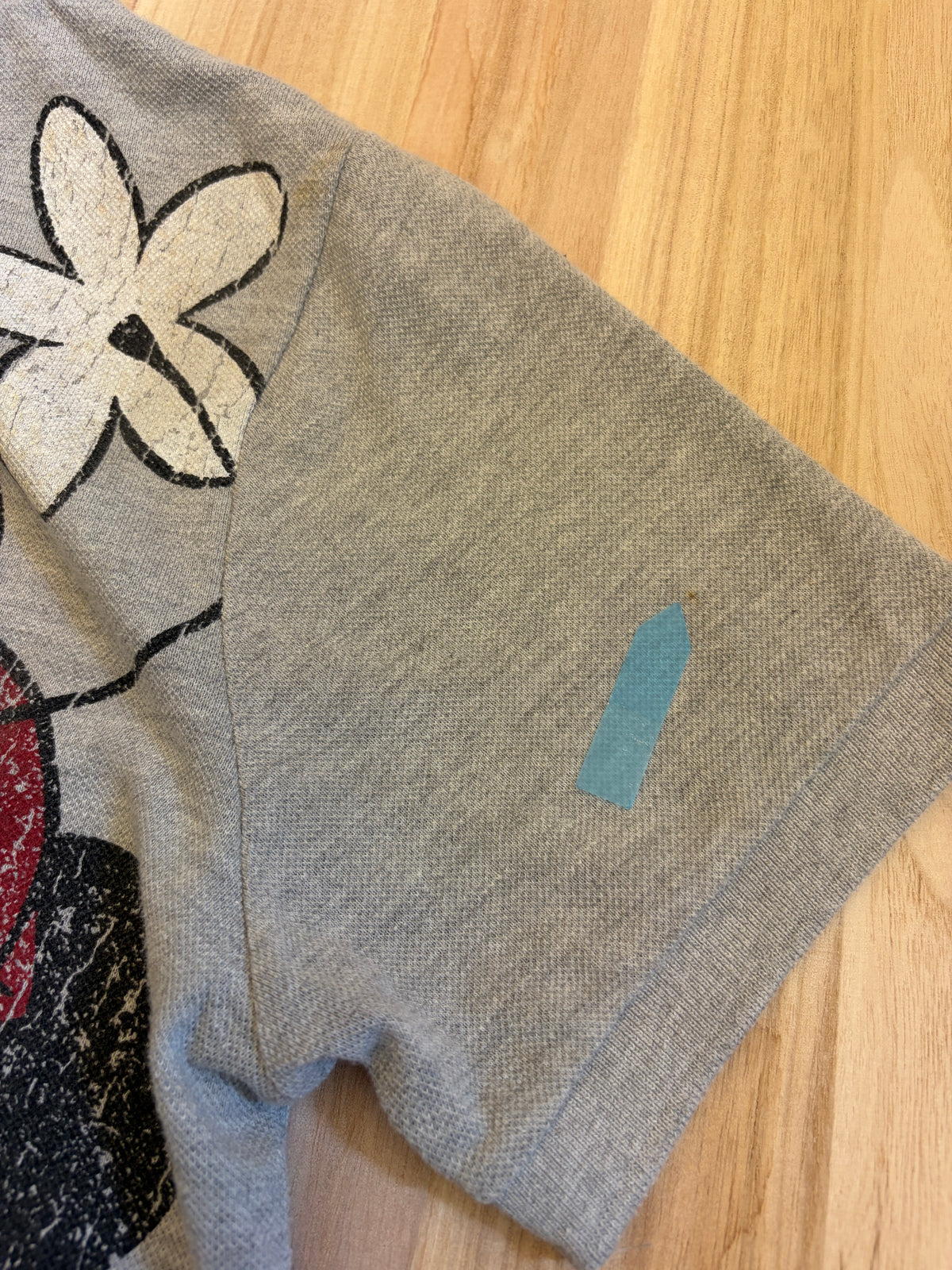 Close-up view of a grey Disney Minnie Mouse polo shirt sleeve cuff showing a small spot stain defect indicated by a blue sticker.