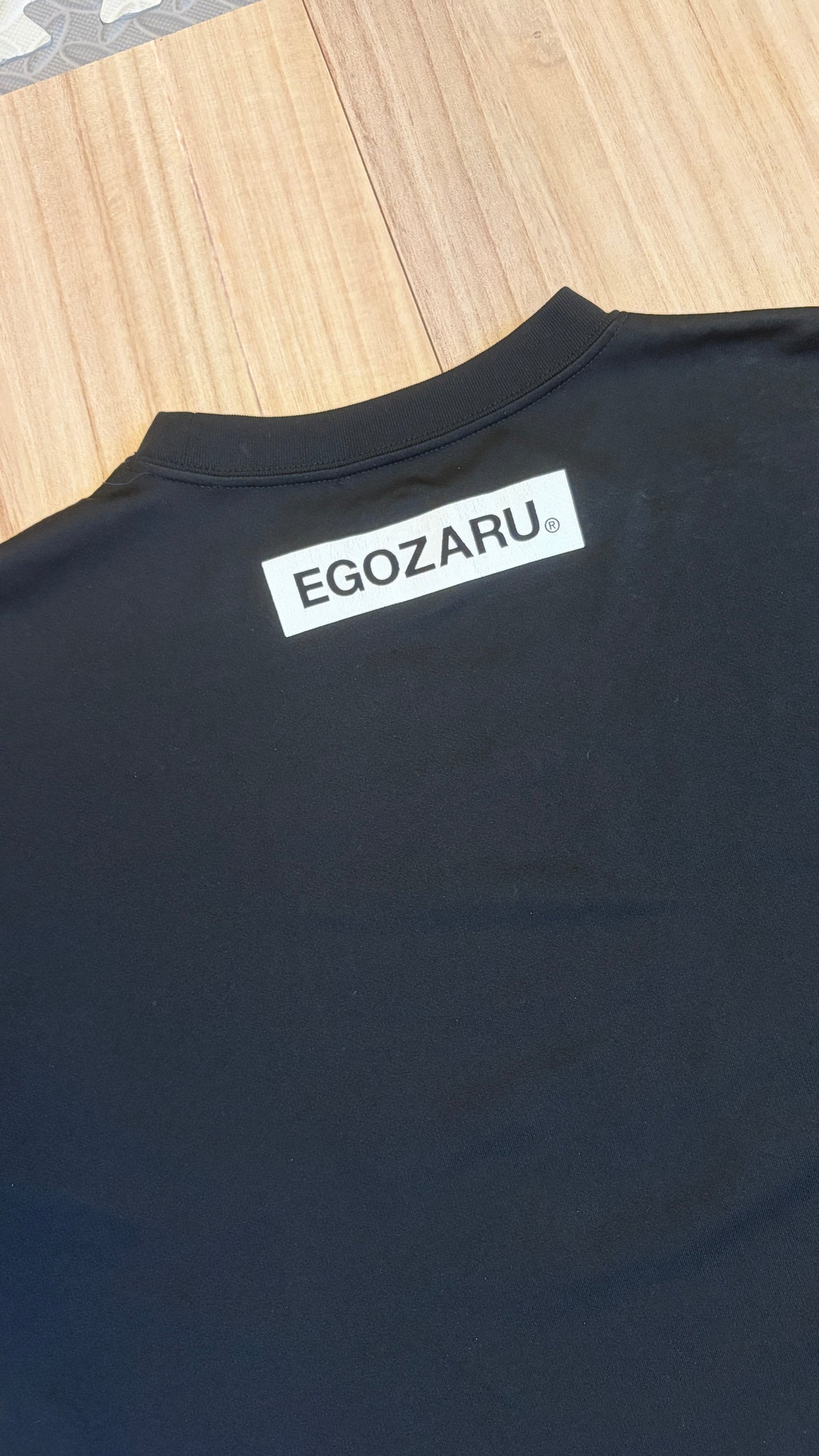 Close-up view of the upper back collar of a black Egozaru athletic t-shirt featuring a white rectangular Egozaru brand logo print