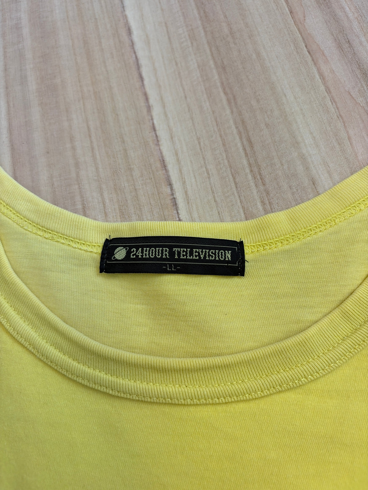 Close-up view of the inner neck collar on a yellow t-shirt displaying a black woven brand tag reading 24HOUR TELEVISION in white block text above size LL
