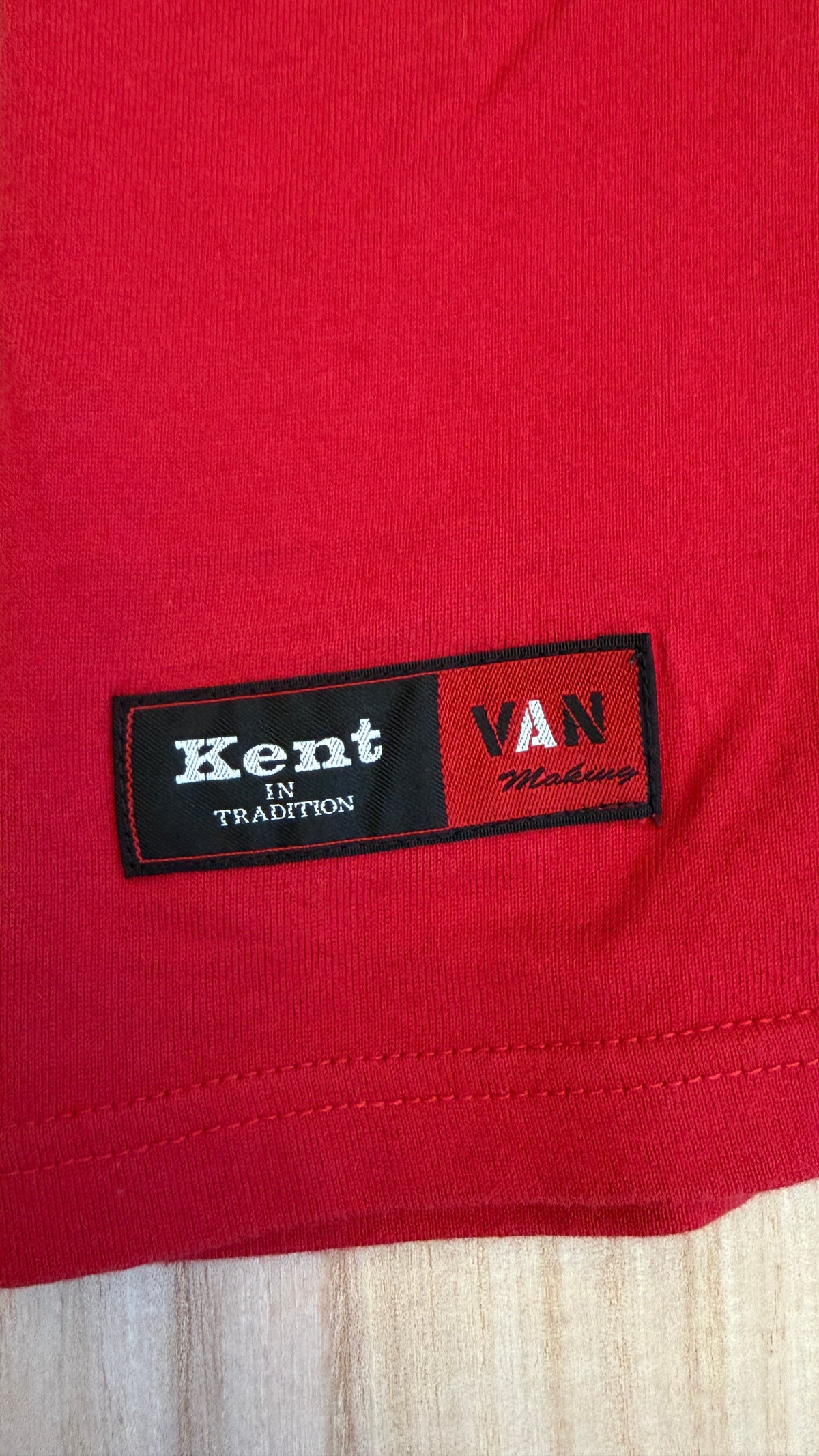 Close-up view of a black and red woven brand patch stitched near the bottom hem of a red t-shirt featuring Kent IN TRADITION and VAN logos