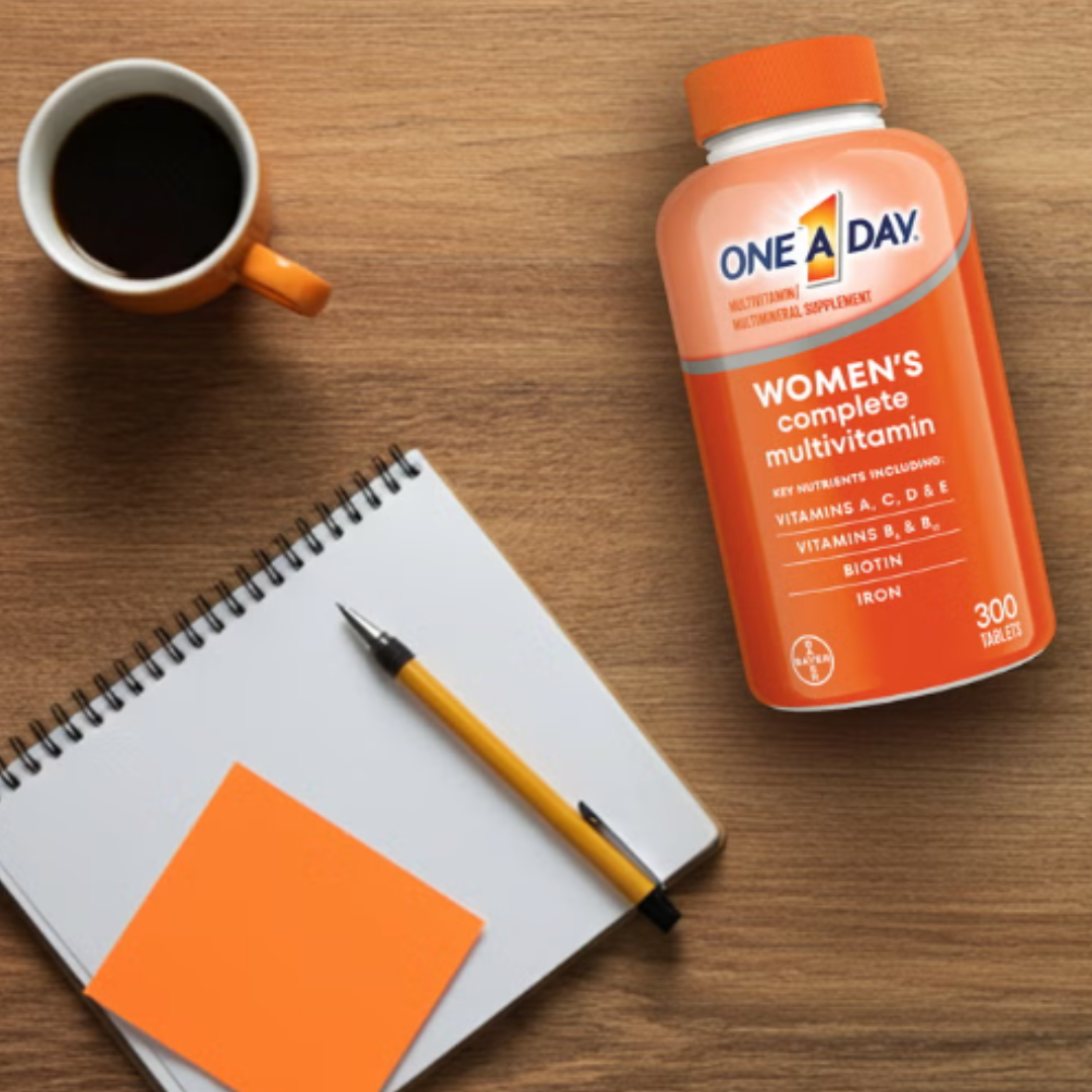 One A Day Women's Complete Multivitamin bottle on a wooden desk next to a coffee cup, notebook, and pen.