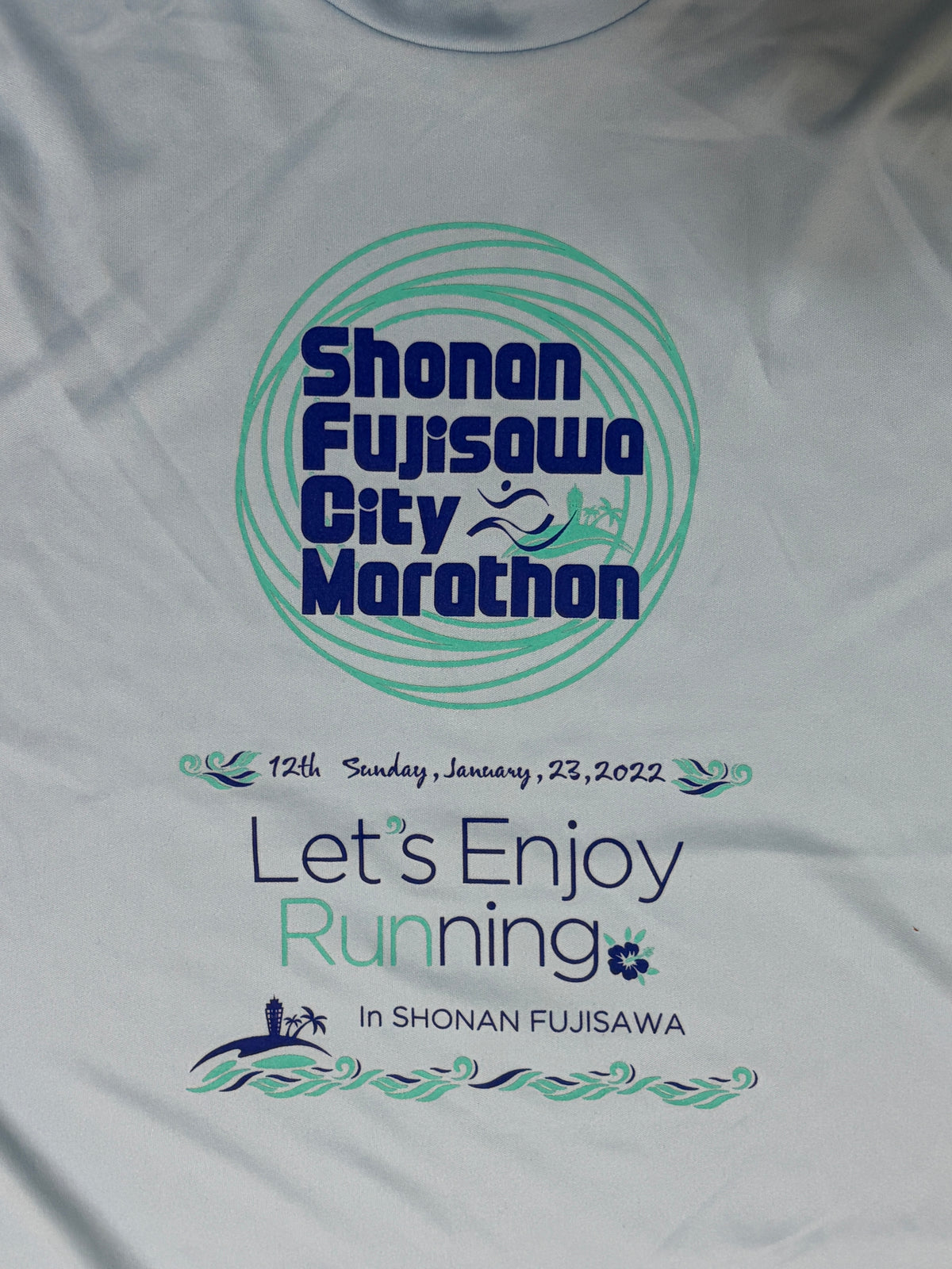 A close-up of a white running shirt featuring a printed graphic for the Shonan Fujisawa City Marathon. The design includes a teal circular logo with dark blue text, the date '12th Sunday, January 23, 2022', and the slogan 'Let's Enjoy Running In SHONAN FUJISAWA' with small running and wave illustrations.