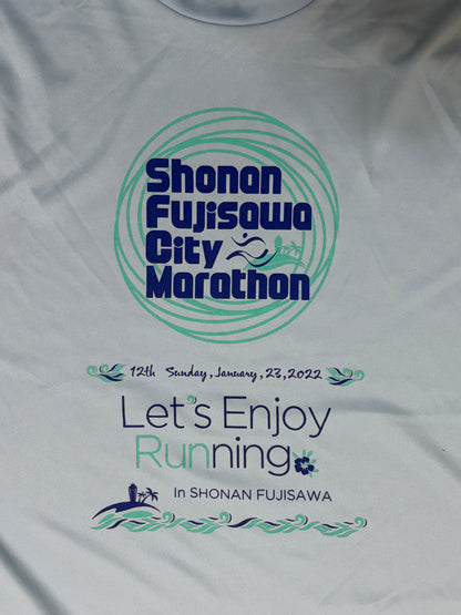 A close-up of a white running shirt featuring a printed graphic for the Shonan Fujisawa City Marathon. The design includes a teal circular logo with dark blue text, the date '12th Sunday, January 23, 2022', and the slogan 'Let's Enjoy Running In SHONAN FUJISAWA' with small running and wave illustrations.