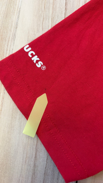 Close-up view of a red Starbucks t-shirt sleeve hem showing a small spot defect indicated by a yellow arrow sticker.