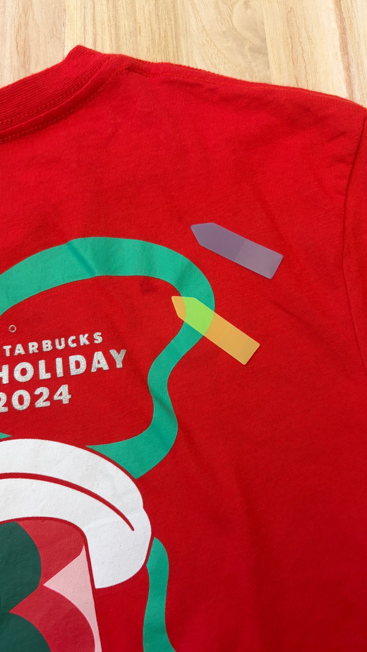 Close-up view of a red Starbucks Holiday 2024 t-shirt showing slight stain defects marked with arrow stickers on the fabric.