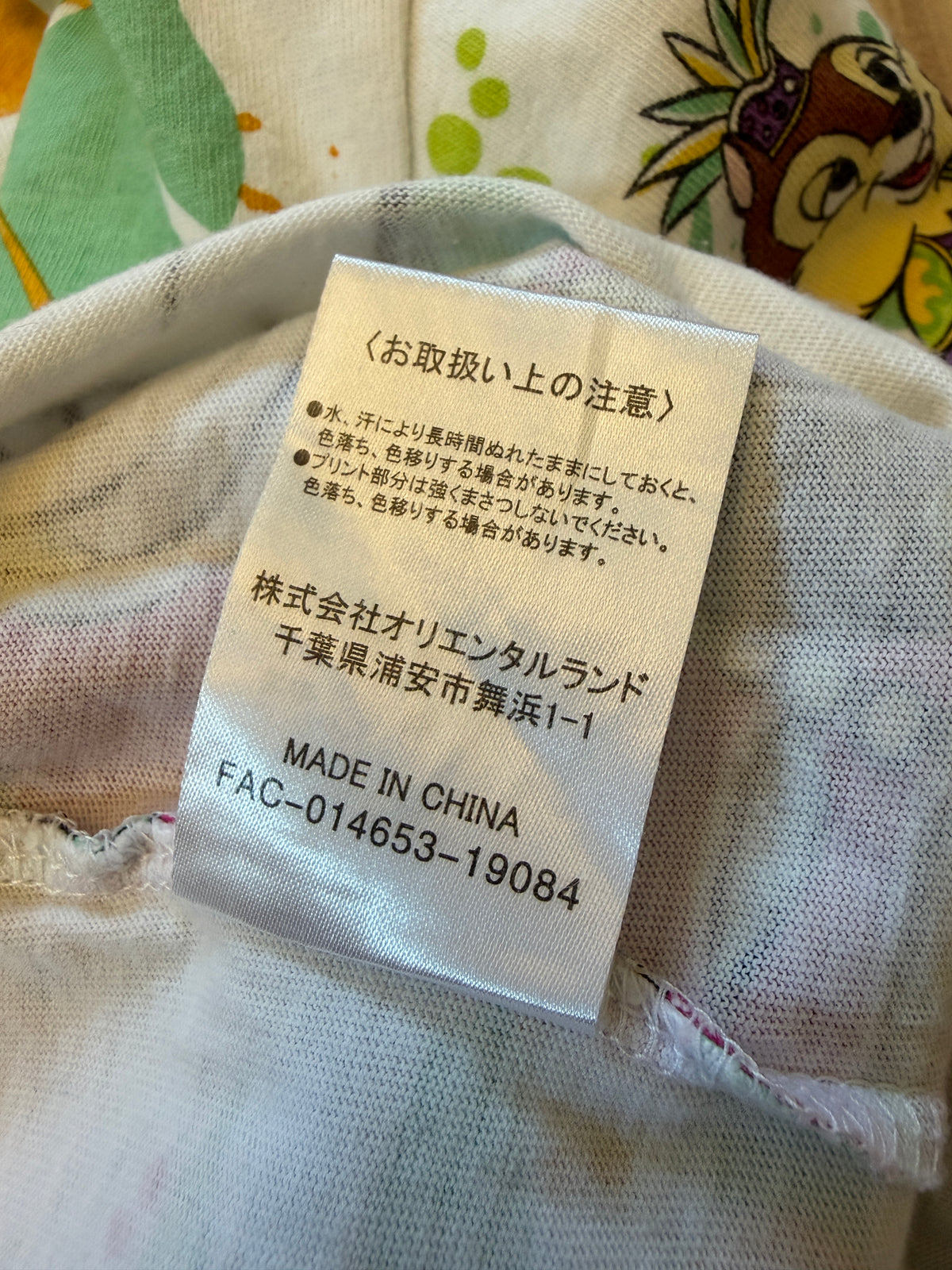 Close-up view of the reverse side of an inner white fabric care tag displaying Japanese handling instructions, The Oriental Land Co., Ltd. details, Made in China, and FAC-014653-19084 manufacturing code