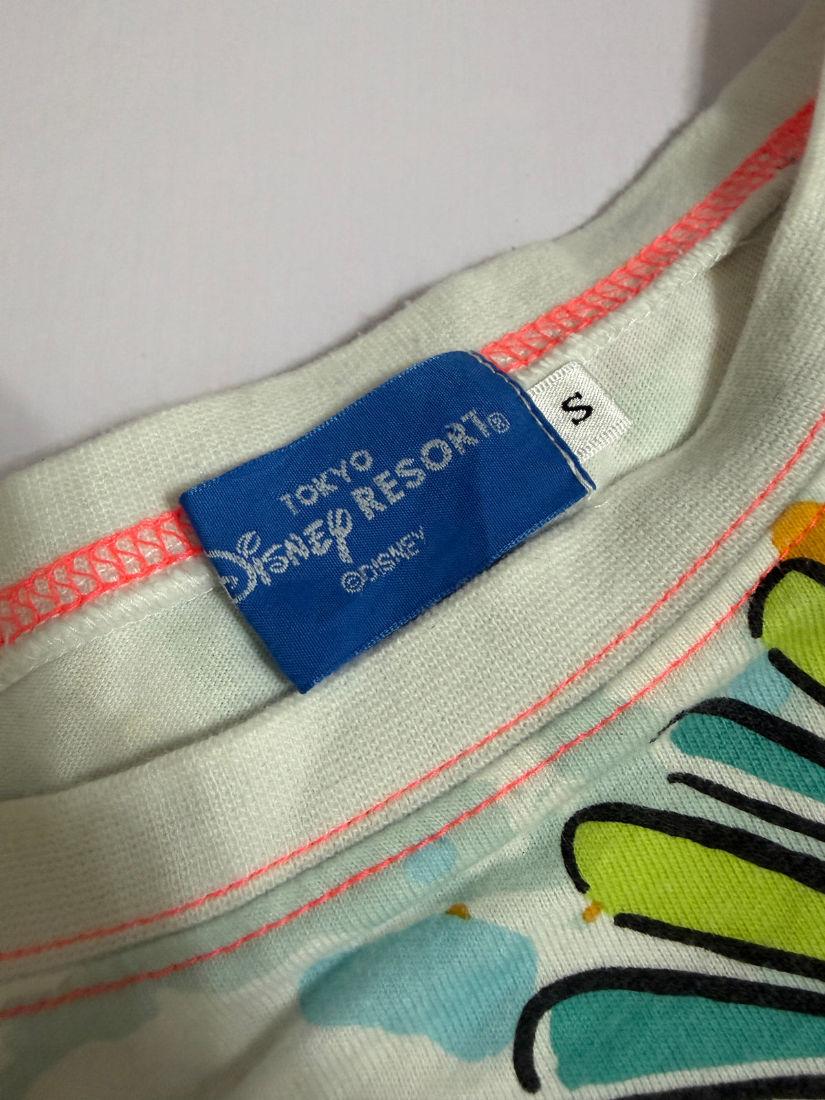Close-up detail of the blue Tokyo Disney Resort neck label and size S tag on the white all-over print t-shirt with pink collar stitching.