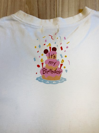 Upper back view of a white Tokyo Disney Resort graphic t-shirt featuring an illustration of a pink Mickey-themed birthday cake surrounded by colorful confetti and text reading It's my Birthday