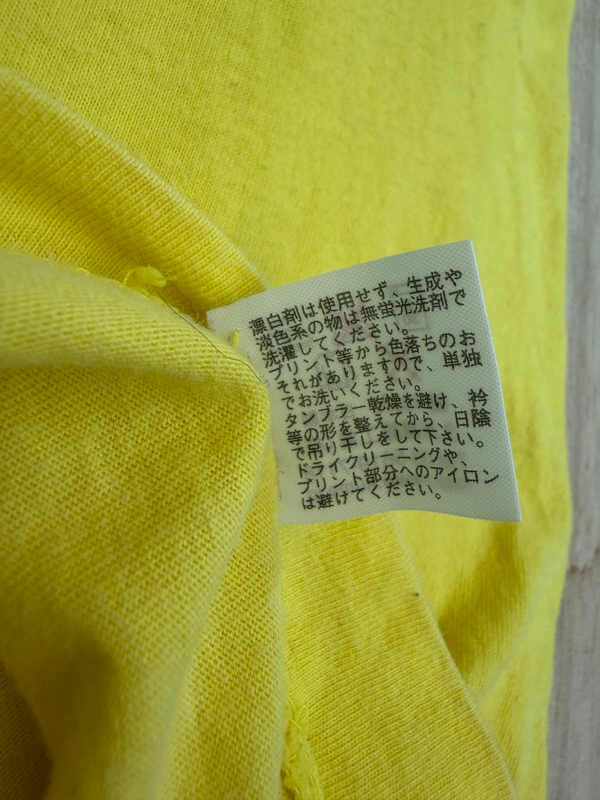 Close-up view of the reverse side of an inner white fabric care tag on a yellow t-shirt displaying Japanese text instructions for washing, drying, and ironing