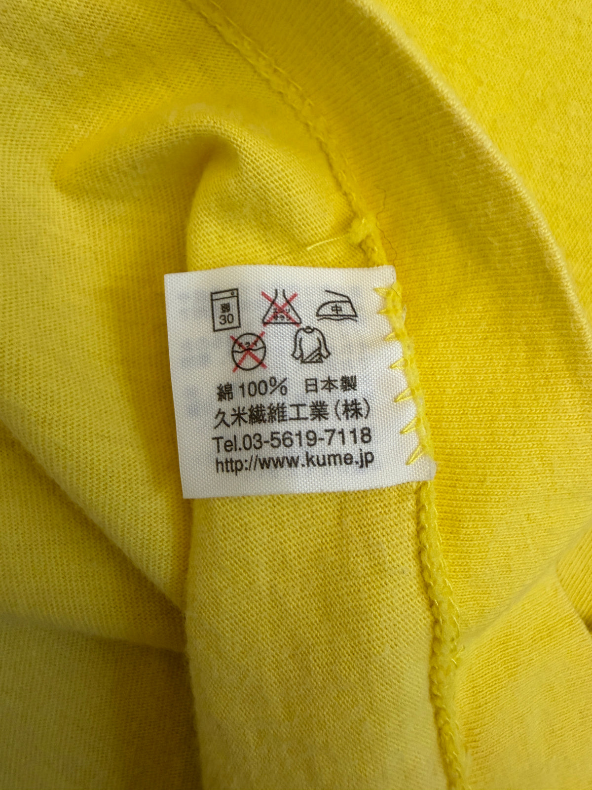 Close-up view of the inner white fabric tag on a yellow t-shirt showing Japanese care symbols, 100% cotton composition, Made in Japan, and Kume Sen-i Co., Ltd. manufacturer details