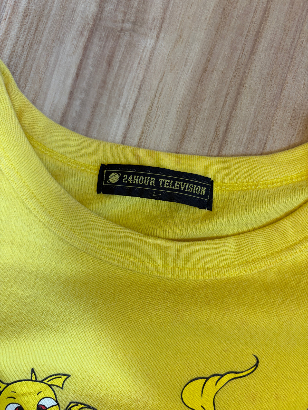 Close-up view of the inner neck collar on a yellow t-shirt displaying a black woven brand label reading 24HOUR TELEVISION in white block text above size L