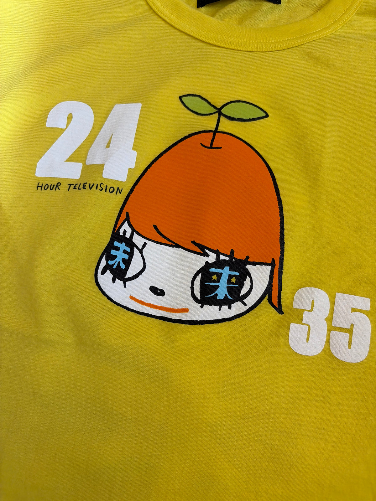Front view of a bright yellow Yoshitomo Nara x 24 Hour Television 35th charity t-shirt featuring a printed character illustration wearing an orange hat with leaf sprout, Kanji characters in the eyes, and numbers 24 and 35